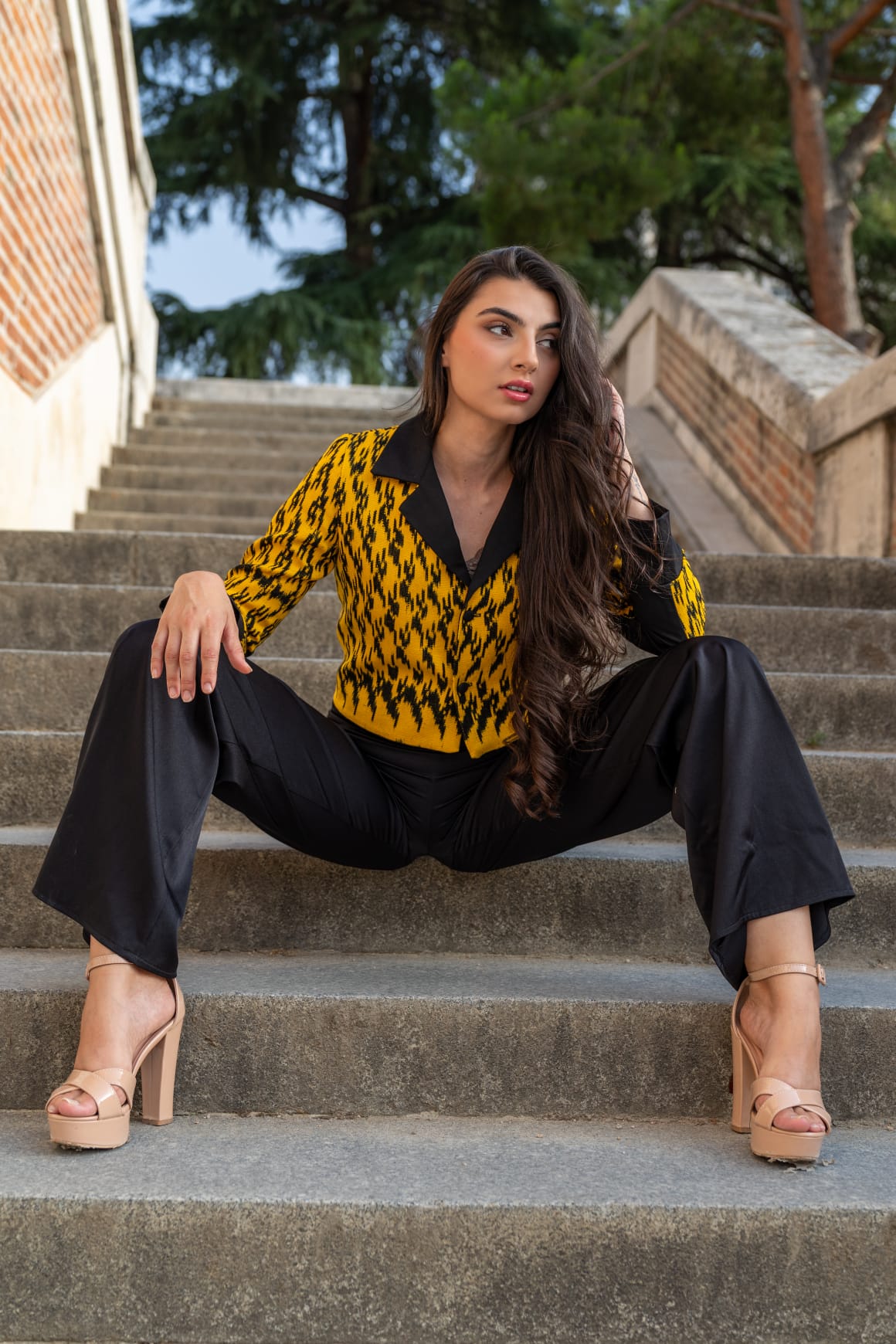 Torera ikat amarillo azafrán y negro con pantalón negro fluido y sandalias nude, modelo sentada en escalinata de piedra