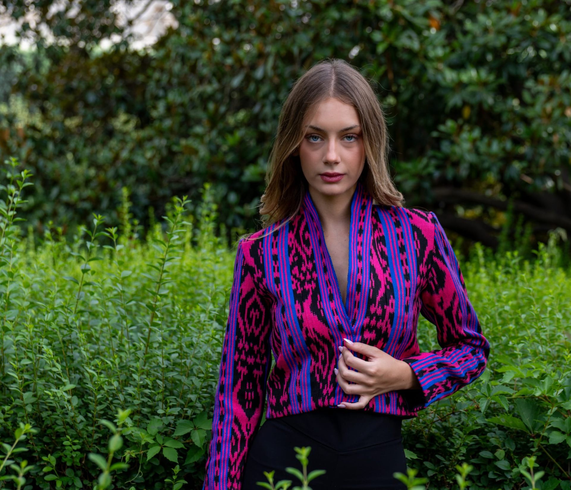 Torera ikat fucsia, azul eléctrico y negro con escote en V, retrato de modelo rubia entre setos verdes