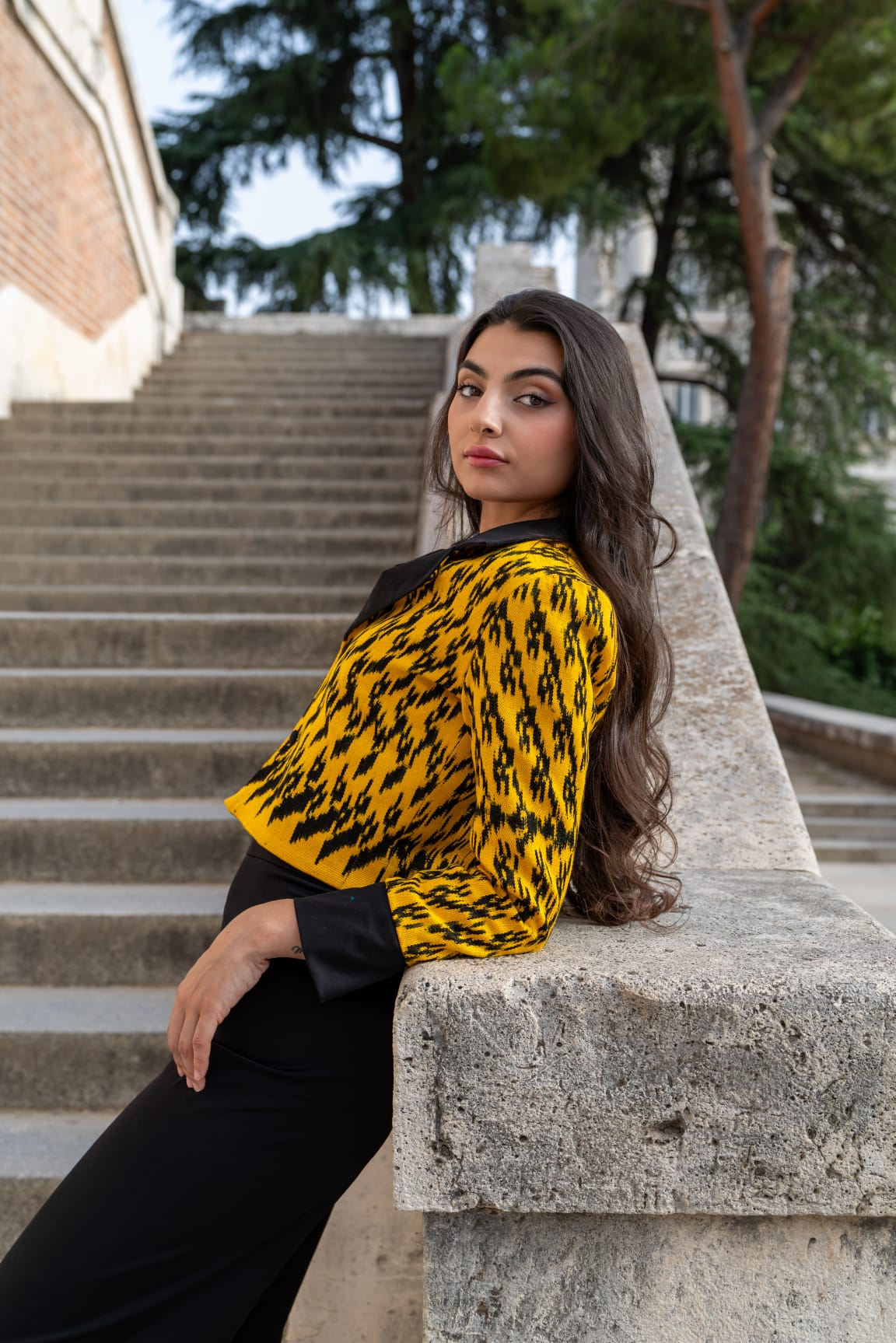 Torera ikat amarillo azafrán y negro combinada con pantalón negro, modelo apoyada en balaustrada de piedra junto a escalinata