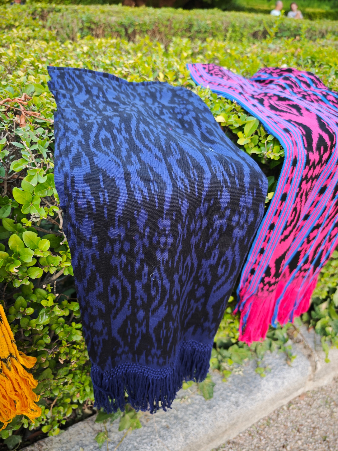 Bufanda ikat azul marino y negro con flecos anudados, apoyada sobre seto verde