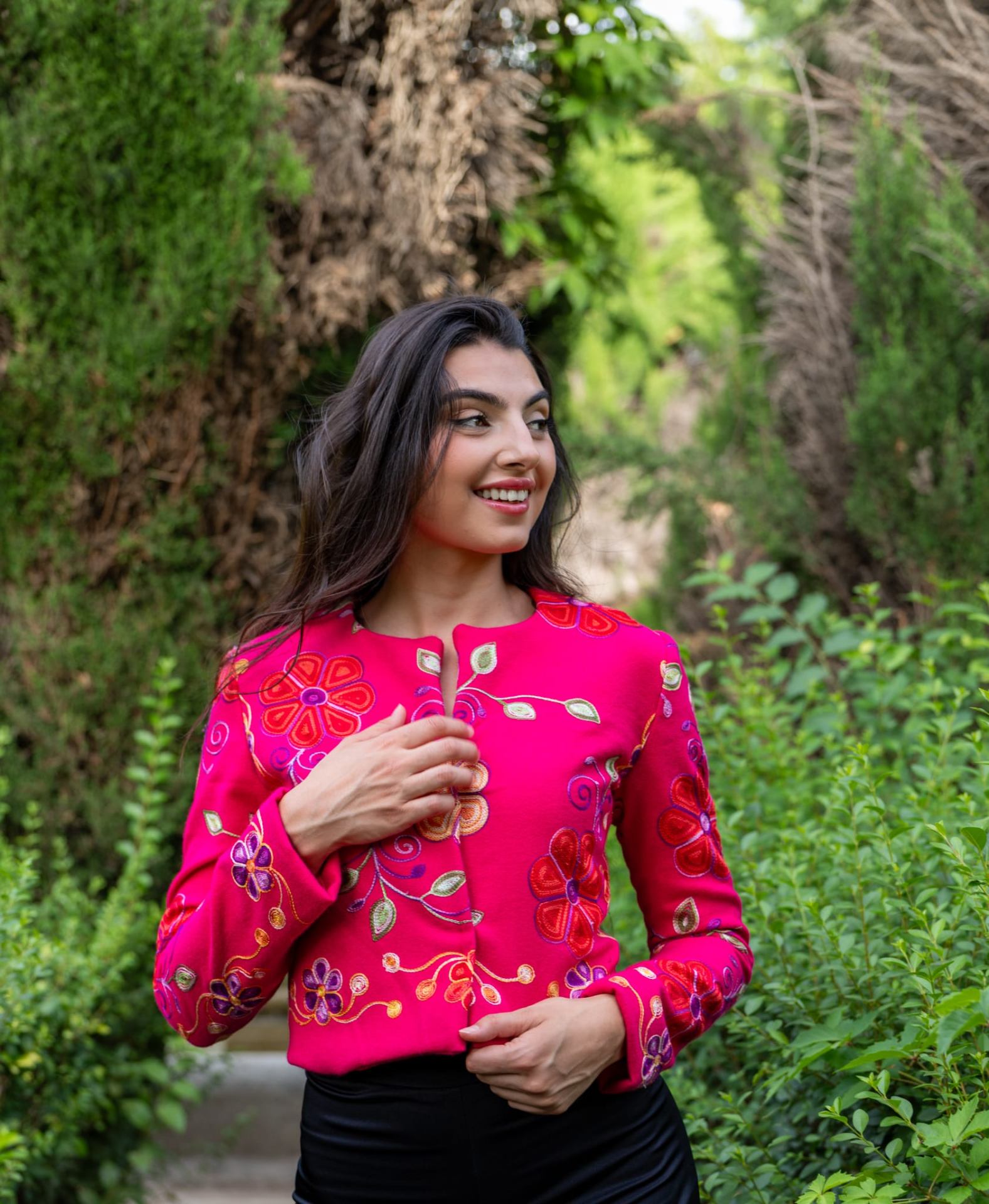 Torera fucsia bordada con flores rojas y moradas, modelo abrochándose la prenda en jardín arbolado