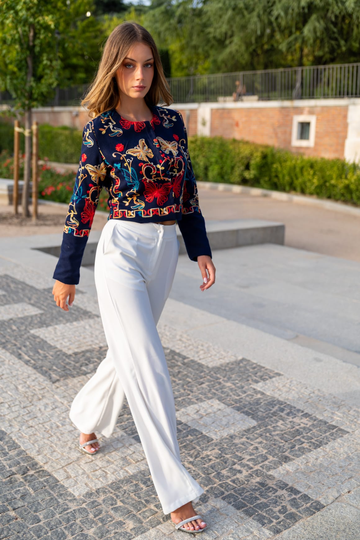 Torera azul marino bordada con mariposas rojas, amarillas y turquesa, combinada con pantalón blanco caminando por adoquín en Madrid