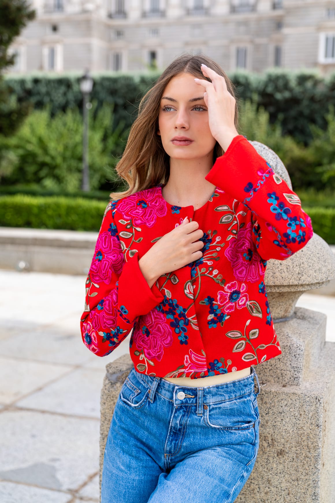 Torera roja bordada con rosas fucsia y flores azules, modelo apoyada en balaustrada de piedra con vaqueros